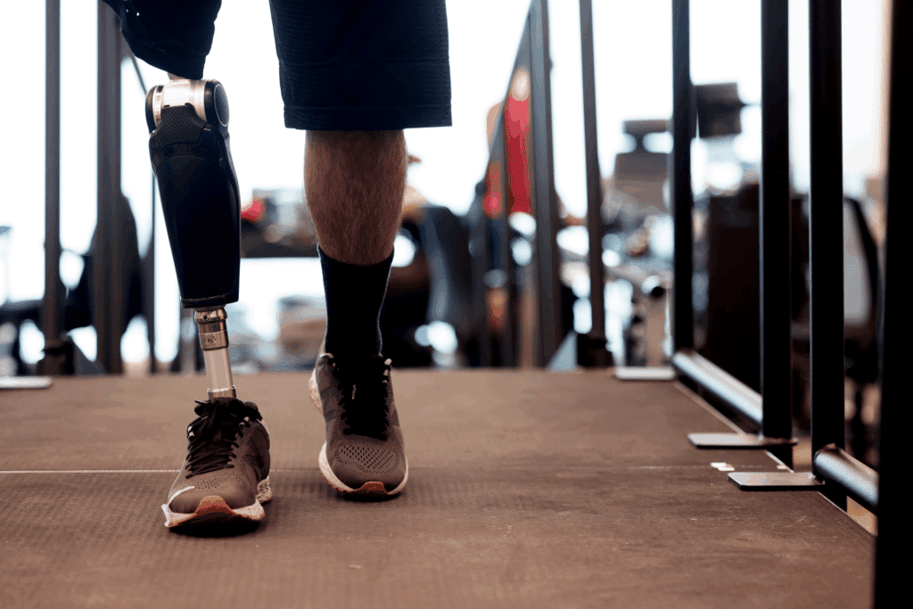 The walking study participant shifts weight onto the prosthesis, which is silver and black. The housing for the motor and electronics are roughly the shape of the knee and calf, narrowing to a metal post above the athletic shoe covering the foot.